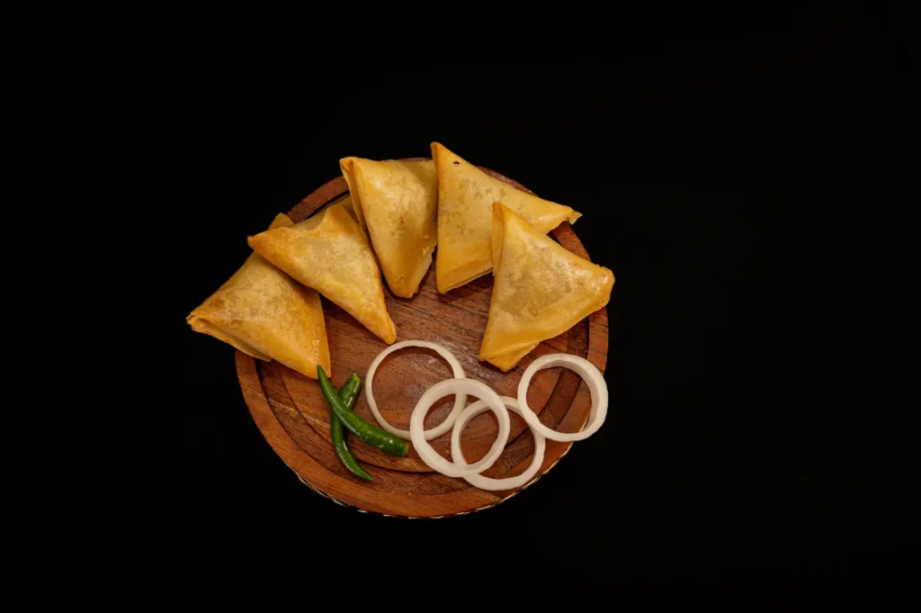 Crispy golden samosas served on a wooden plate with onion rings and green chilies against a black background.
