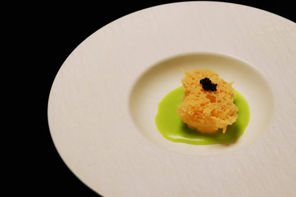 A white plate showcases a sophisticated dish featuring a small, airy pastry atop vibrant green sauce, crowned with a tiny dollop of black caviar.