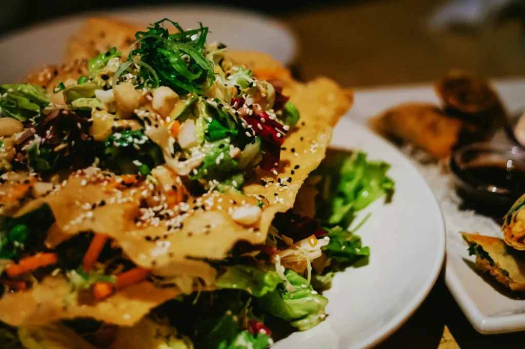 A vibrant salad features mixed greens, nuts, and sesame seeds layered with crispy chips, creating a fresh and crunchy texture. A side dish is blurred in the background.