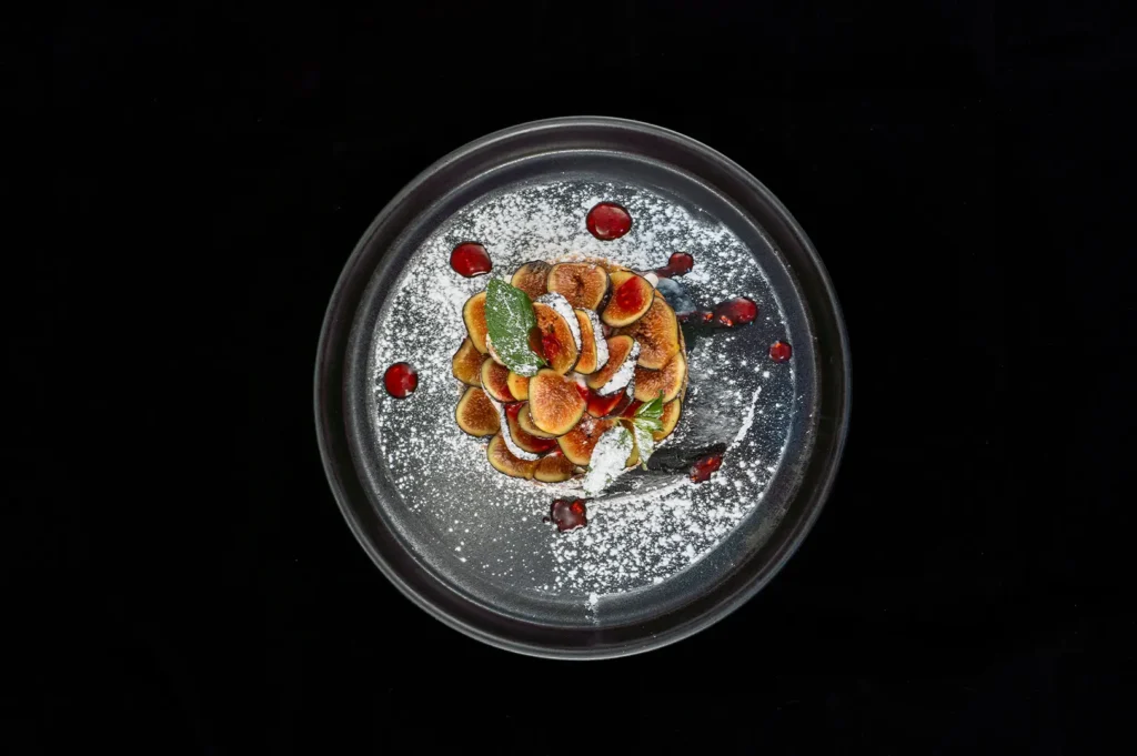 Overhead shot of a gourmet fig dessert garnished with powdered sugar, fresh mint, and berry reduction on a dark plate.