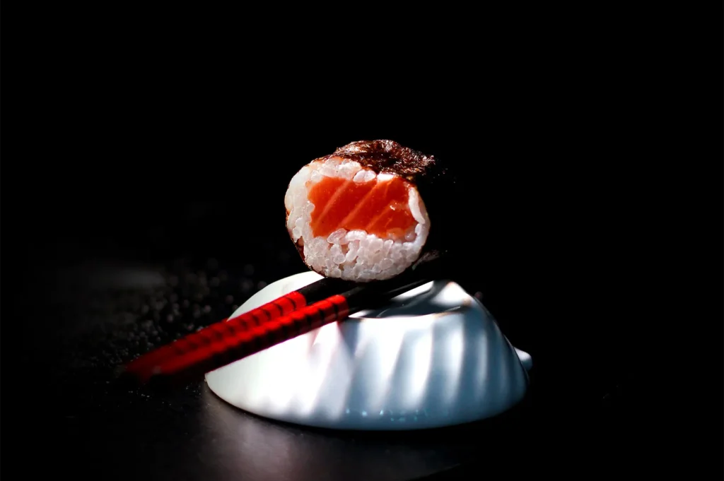 Single piece of salmon maki sushi balanced on red and black chopsticks against a black background.