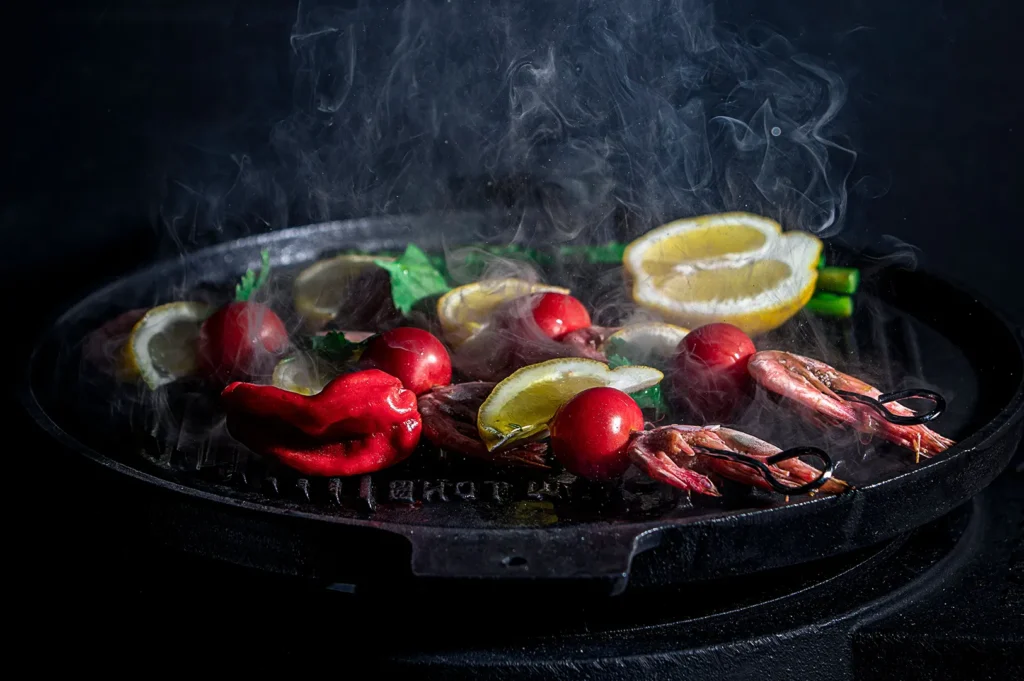 Moody black food photography of sizzling grilled seafood, lemons, and herbs with rising steam on a dark skillet.