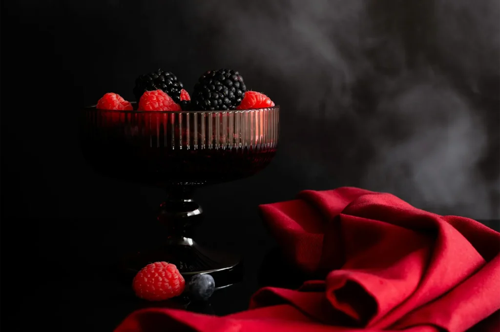 Fresh raspberries and blackberries in a dark glass pedestal bowl on a black surface with a red cloth.