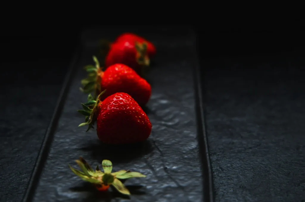 Four ripe strawberries on a dark, textured slate board, arranged in a line. The berries' vibrant red contrasts with the dark background, creating a fresh, elegant tone.