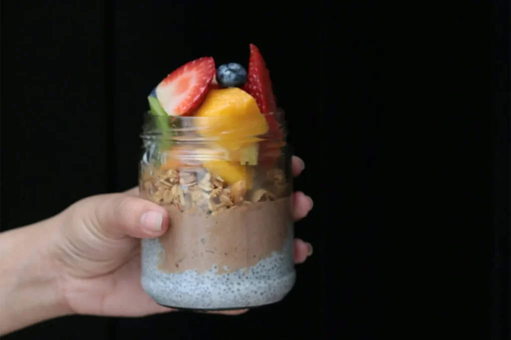 Hand holding a jar of chocolate chia seed pudding topped with fresh strawberries, mango, and blueberries.