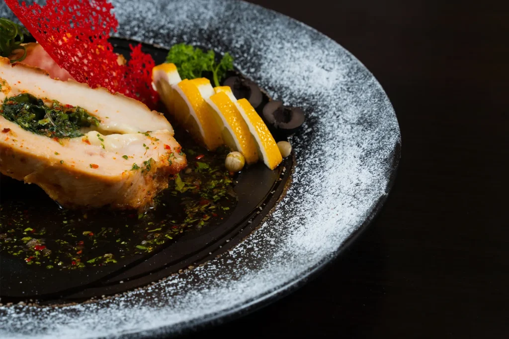 Gourmet stuffed chicken breast with sauce and lemon slices on a dark plate.