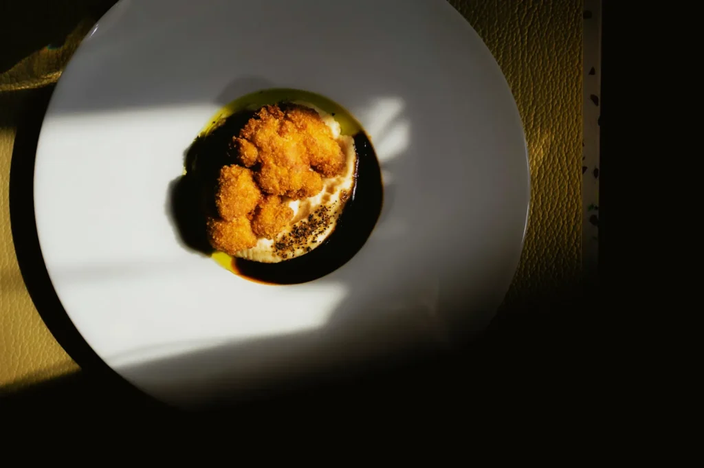 A dish on a white plate is bathed in dramatic light, showcasing golden brown fried morsels atop a creamy sauce with green oil accents. The setting feels artful and elegant.