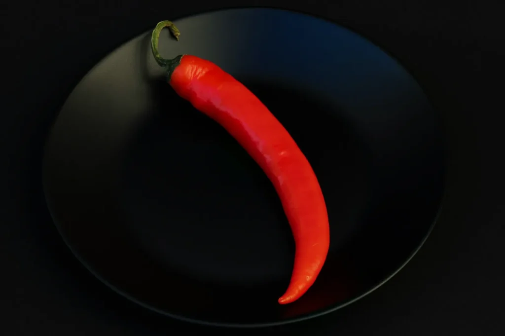A vivid red chili pepper is placed on a black plate against a dark background, creating a striking contrast. The scene conveys simplicity and boldness.