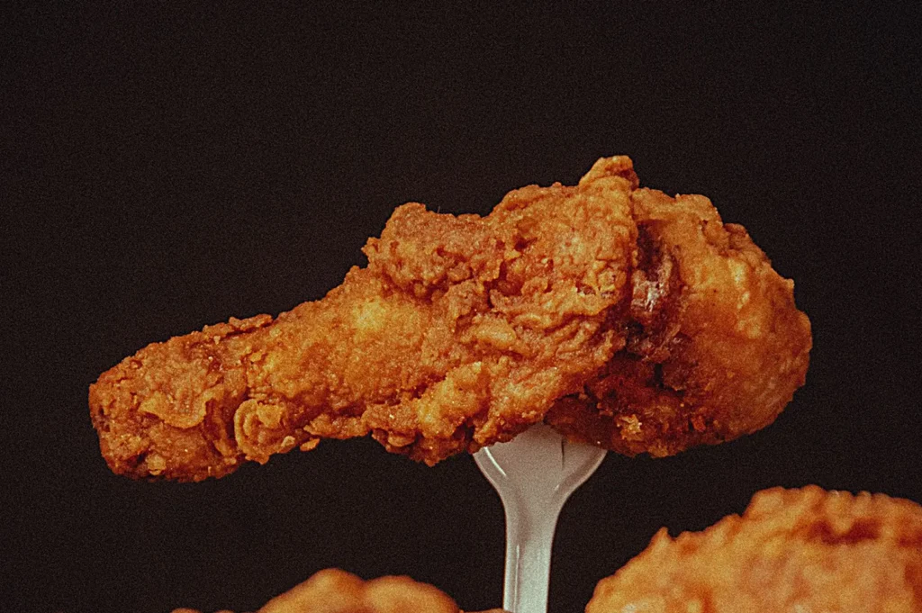 Crispy golden fried chicken drumstick on a fork against a dark background.