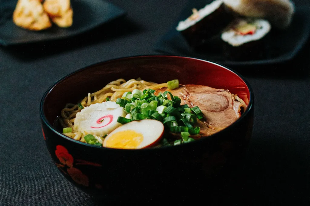 A bowl of Japanese ramen topped with chashu pork, a soft-boiled egg, narutomaki fish cake, and chopped green onions, served on a dark background.