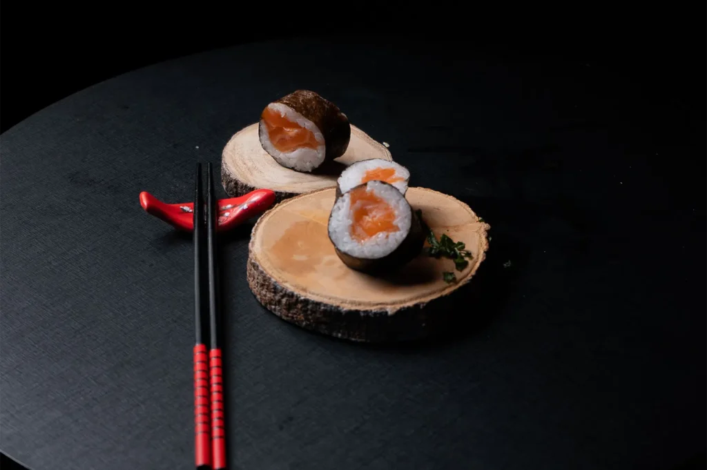 Fine dining salmon sushi rolls on rustic wooden slices with black and red chopsticks, dark moody food photography.