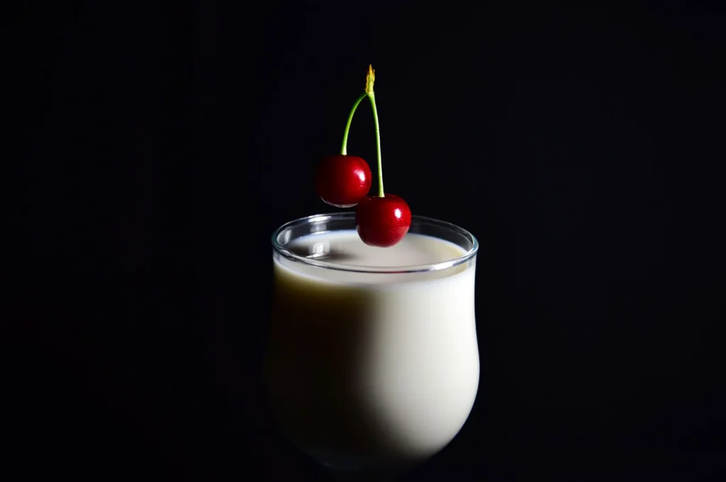 Creamy white milkshake garnished with fresh cherries in a glass against a dark background.