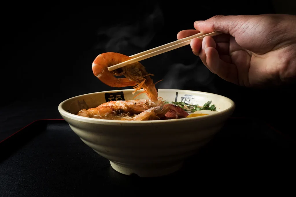 Close-up of steaming shrimp ramen bowl with chopsticks lifting a shrimp against a dark background.
