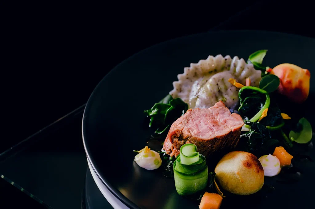 Gourmet plated dish with sliced meat, ravioli, and vegetables on a dark plate.
