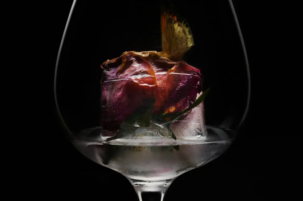 A delicate butterfly sits on a vibrant pink rose encased in a large ice cube, placed inside a clear wine glass, set against a dark background.