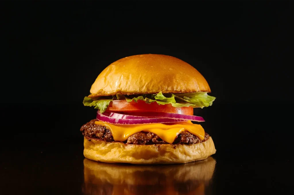 Eye-level shot of a classic cheeseburger with a beef patty, melted cheddar cheese, fresh lettuce, tomato, and red onion on a brioche bun against a black background.