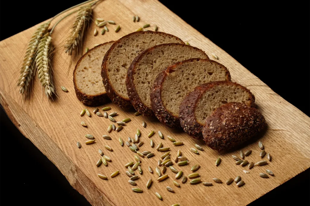 Sliced whole grain bread on a wooden board with wheat stalks and seeds.