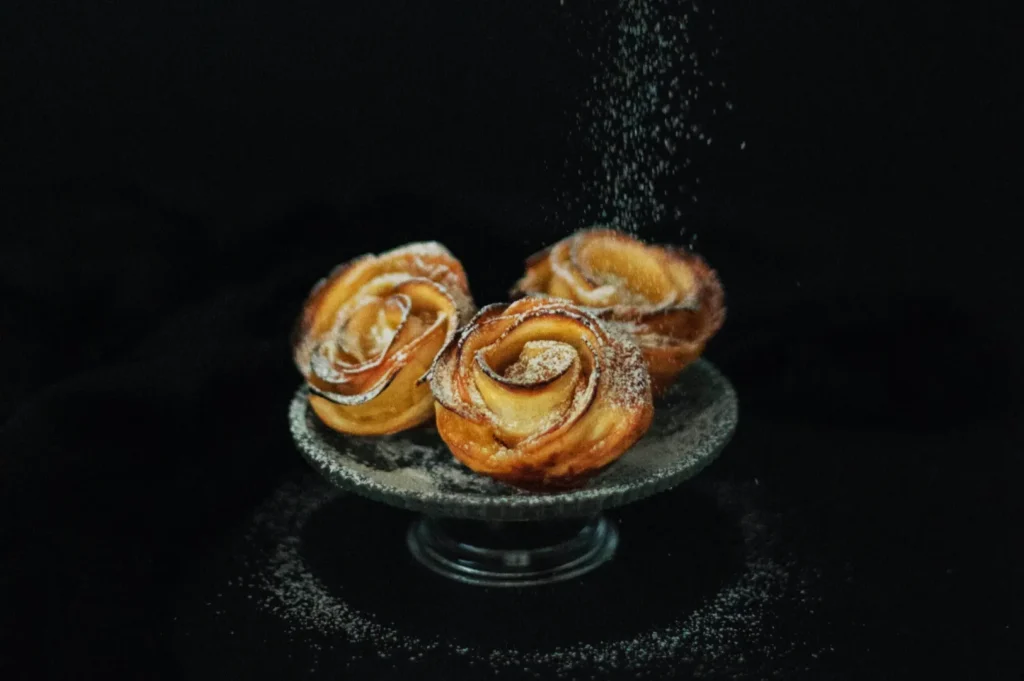 Three rose-shaped pastries dusted with powdered sugar on a pedestal against a dark background, conveying elegance and sweetness.