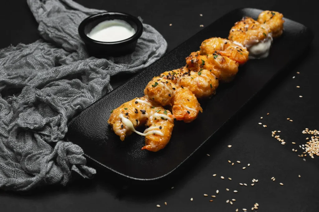 A skewer of crispy golden fried shrimp drizzled with sauce and topped with black sesame seeds on a black plate with a side of dipping sauce.