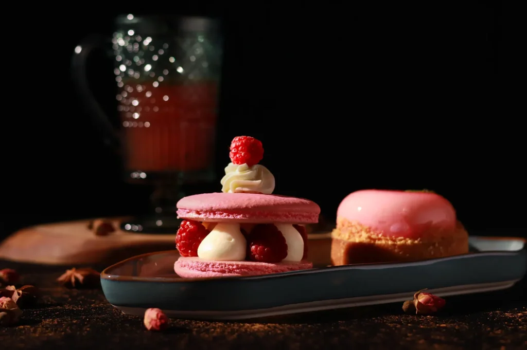 Dramatic low-key food photography featuring pink raspberry desserts and a drink, utilizing side lighting to emphasize texture and mood on a dark background.