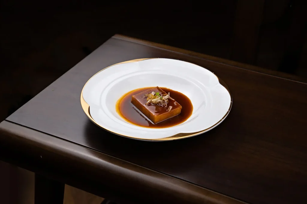 Smooth and silky tofu dish served in savory sauce on a white plate with gold trim.