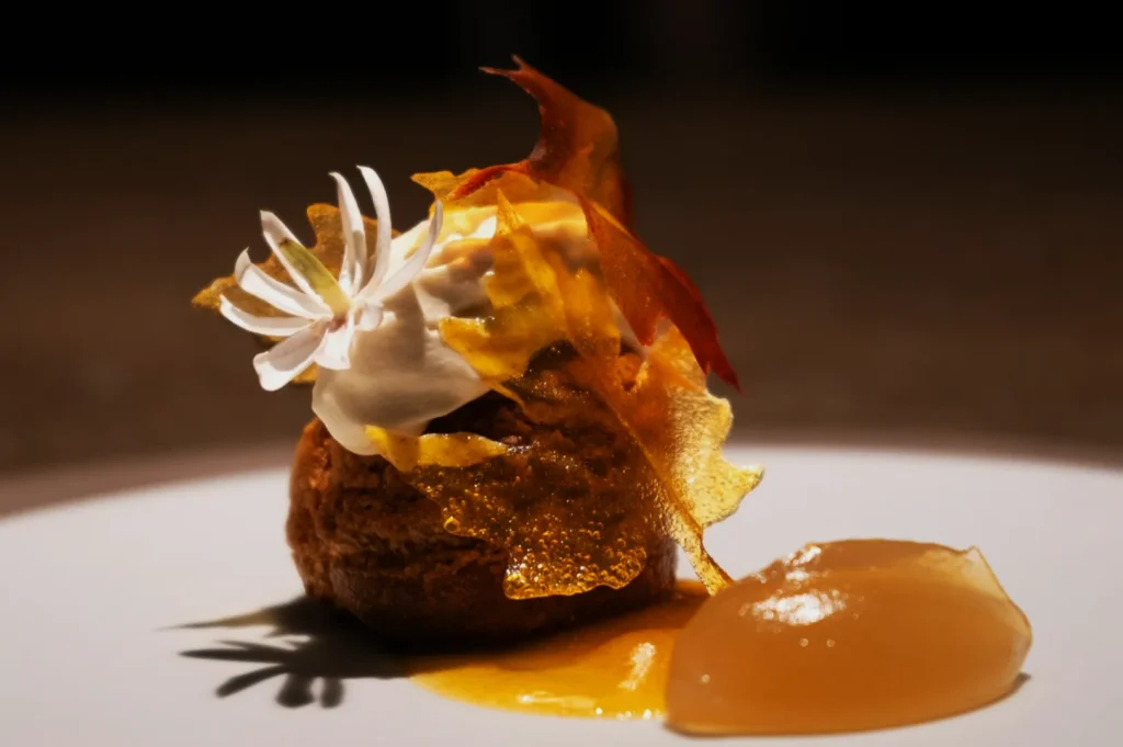 An elegant dessert on a white plate features a small pastry topped with whipped cream, a white flower decoration, and caramelized sugar shards, with a dollop of translucent pear jelly beside it.