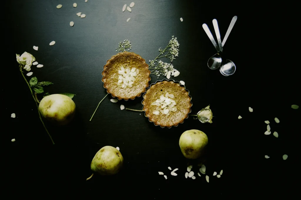 Two pear tarts with almond slices sit on a dark surface surrounded by whole pears, scattered petals, a white flower, and three spoons, evoking an elegant, rustic vibe.