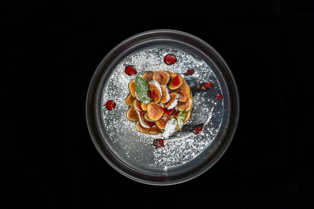 Dark moody food photography of a plated fig tart on a black background, garnished with fresh green mint leaves, red coulis dots, and powdered sugar.