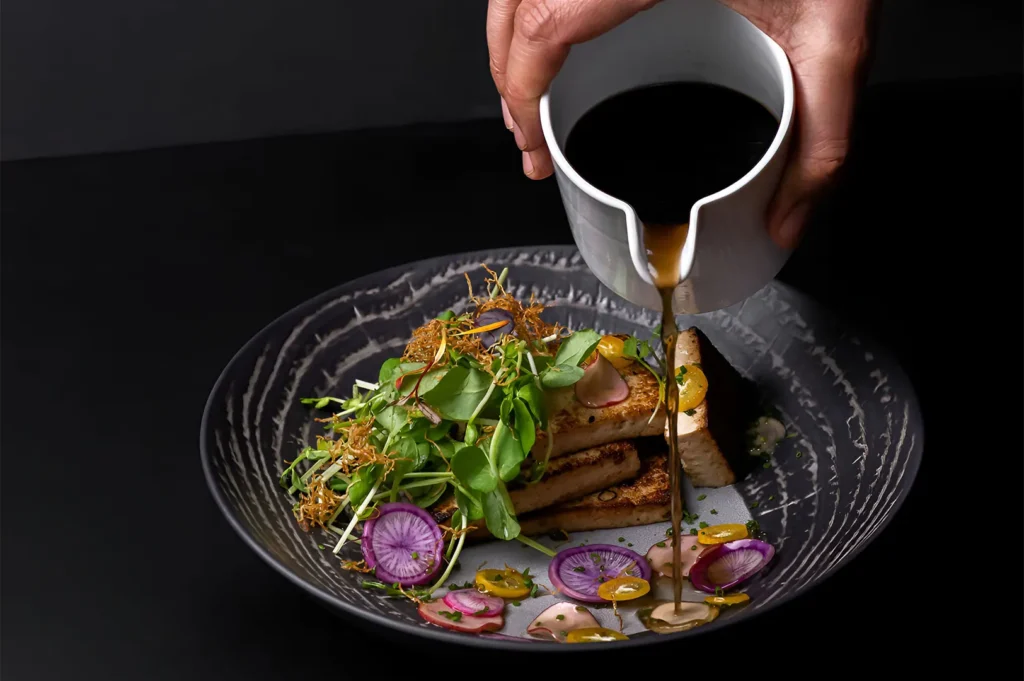 A hand pouring savory sauce over a gourmet dish of pan-seared tofu, fresh microgreens, and sliced radishes on a dark ceramic plate.