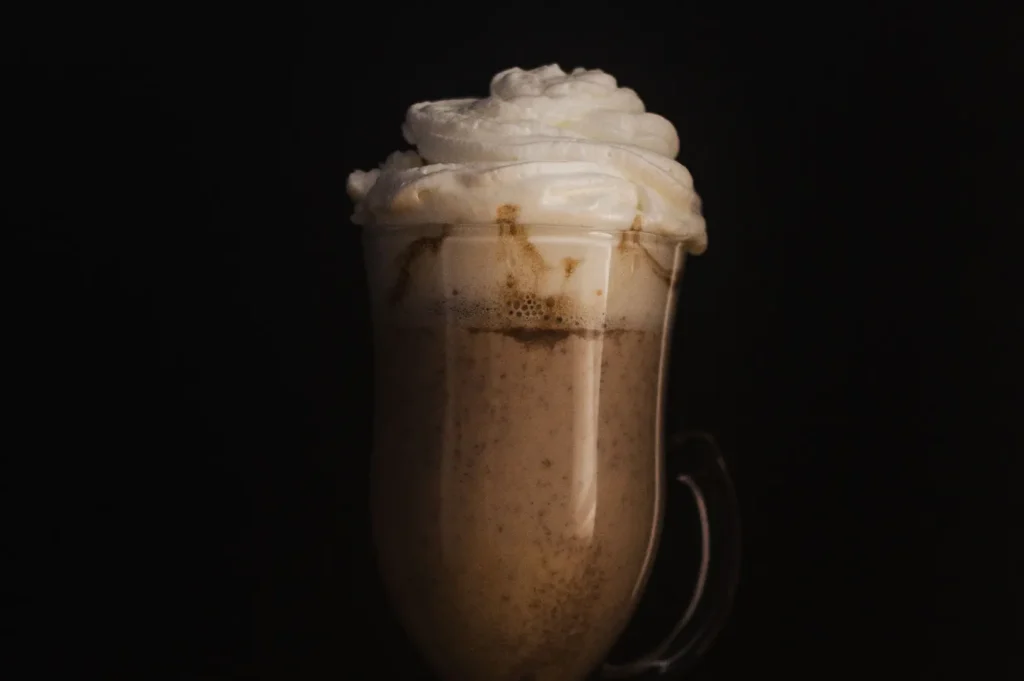 Creamy iced coffee topped with whipped cream in a glass mug against a dark background.