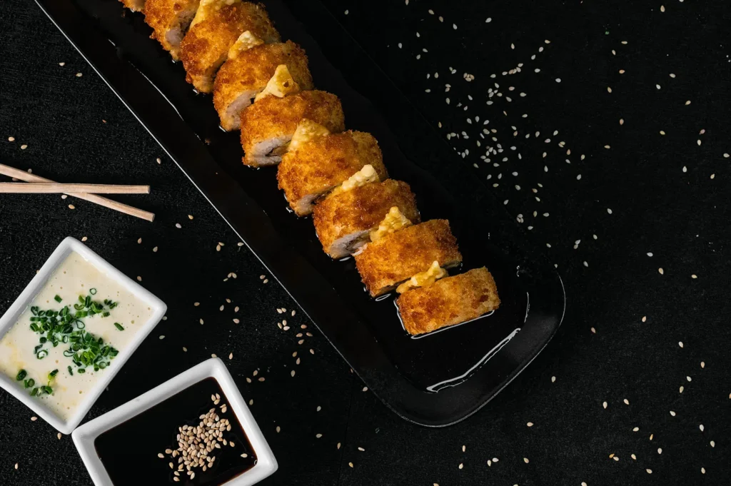 Crispy fried sushi roll on a black plate with dipping sauces.