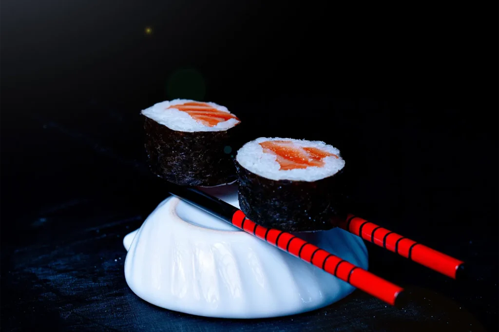 Fresh salmon maki sushi rolls held by red-patterned chopsticks resting on a white ceramic chopstick holder with locked white balance photography.