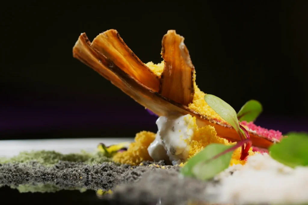 A gourmet dish with a caramelized banana slice, colorful powders, creamy dollops, and microgreens, artfully arranged, creating a vibrant, artistic presentation.