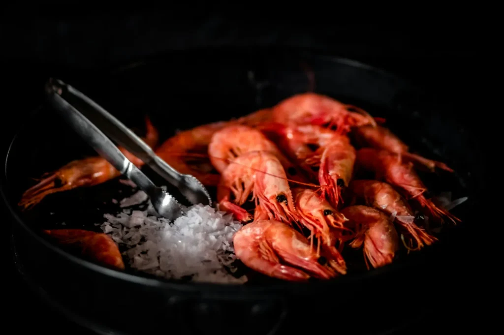 A black pan filled with vibrant pink shrimp and a mound of coarse salt, accompanied by silver tongs. The scene conveys a rustic, appetizing tone.