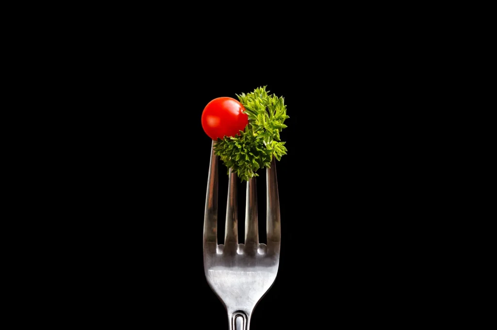 A single cherry tomato and a sprig of fresh green parsley on a silver fork against a black background.