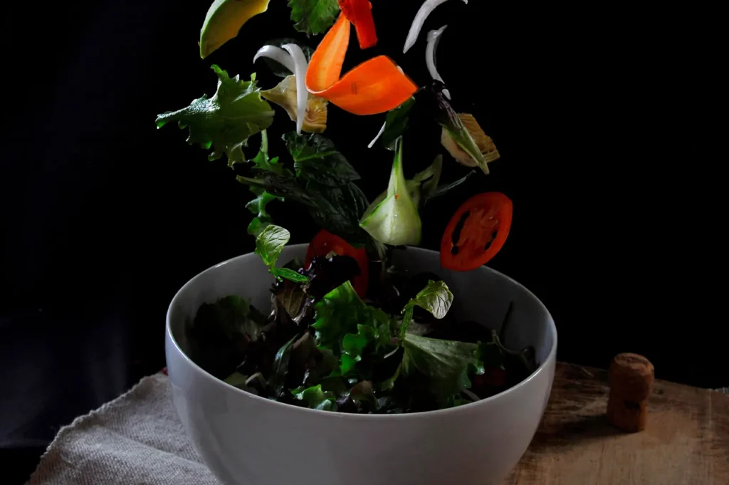 A dark moody food photography of a fresh vegetable salad with vibrant ingredients splashing out of a white bowl against a black background.