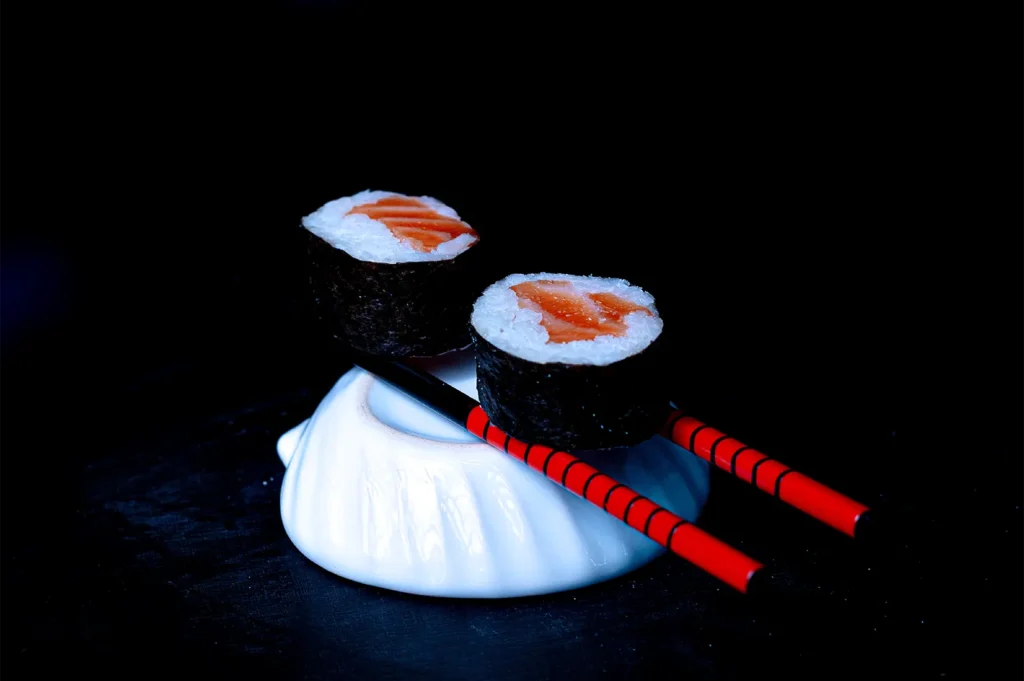 Two pieces of salmon maki sushi resting on red and black chopsticks.