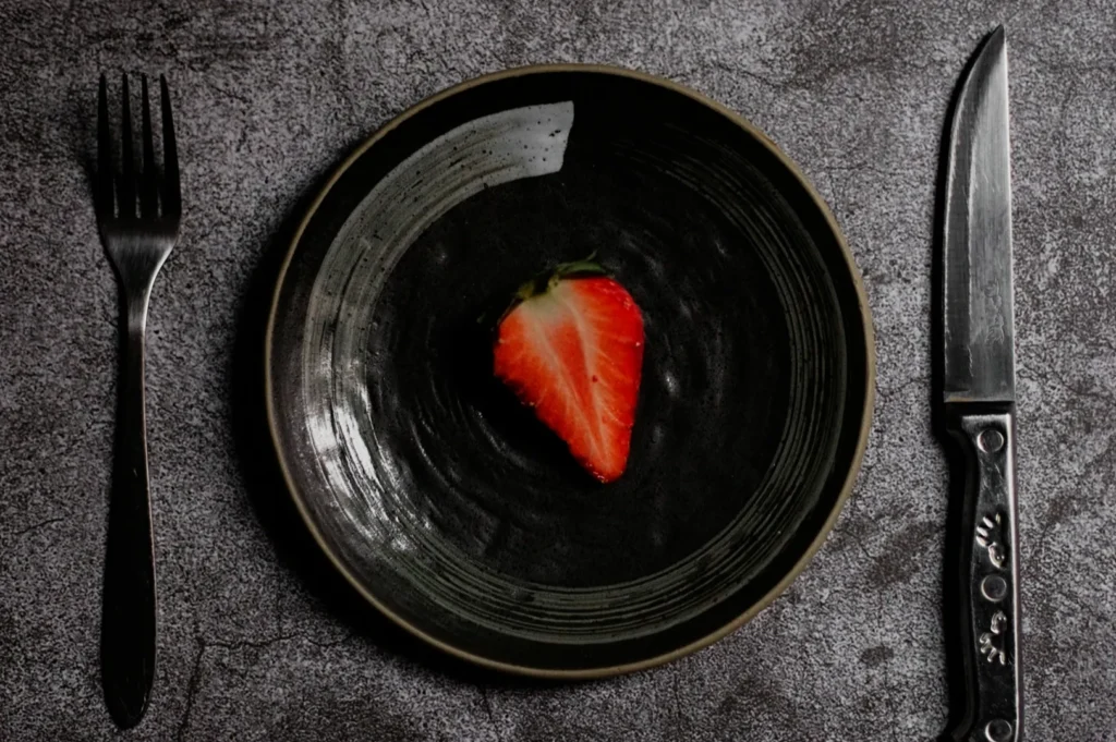 A single strawberry half sits on a dark plate, centered on a textured gray surface. A fork and knife are placed neatly on either side, evoking minimalism.