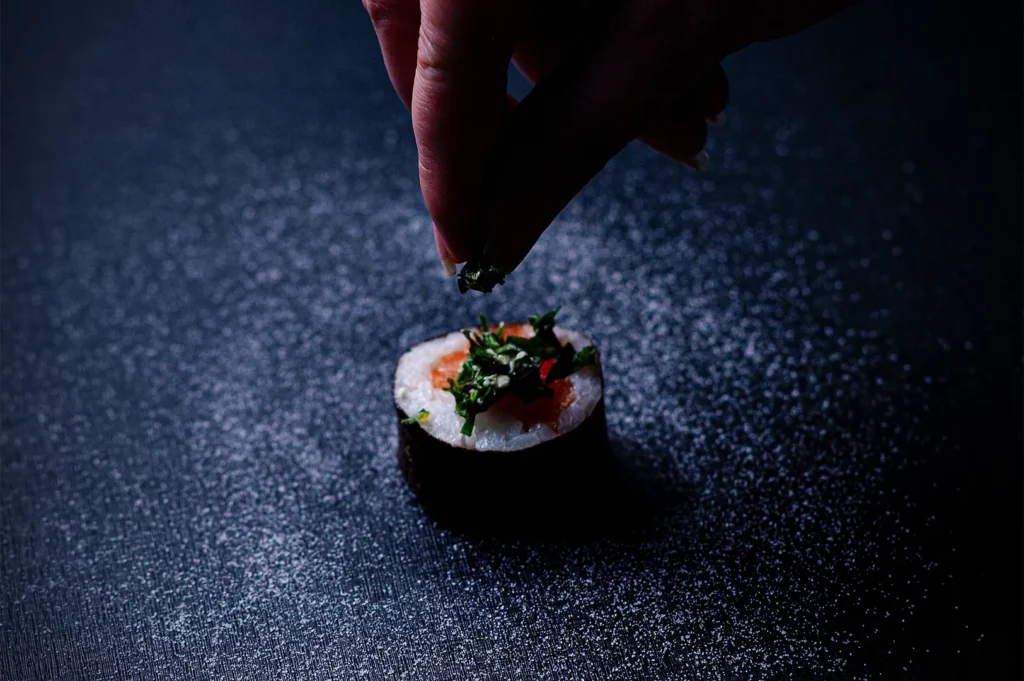 A hand garnishing a single piece of fresh salmon sushi roll on a dark textured background.