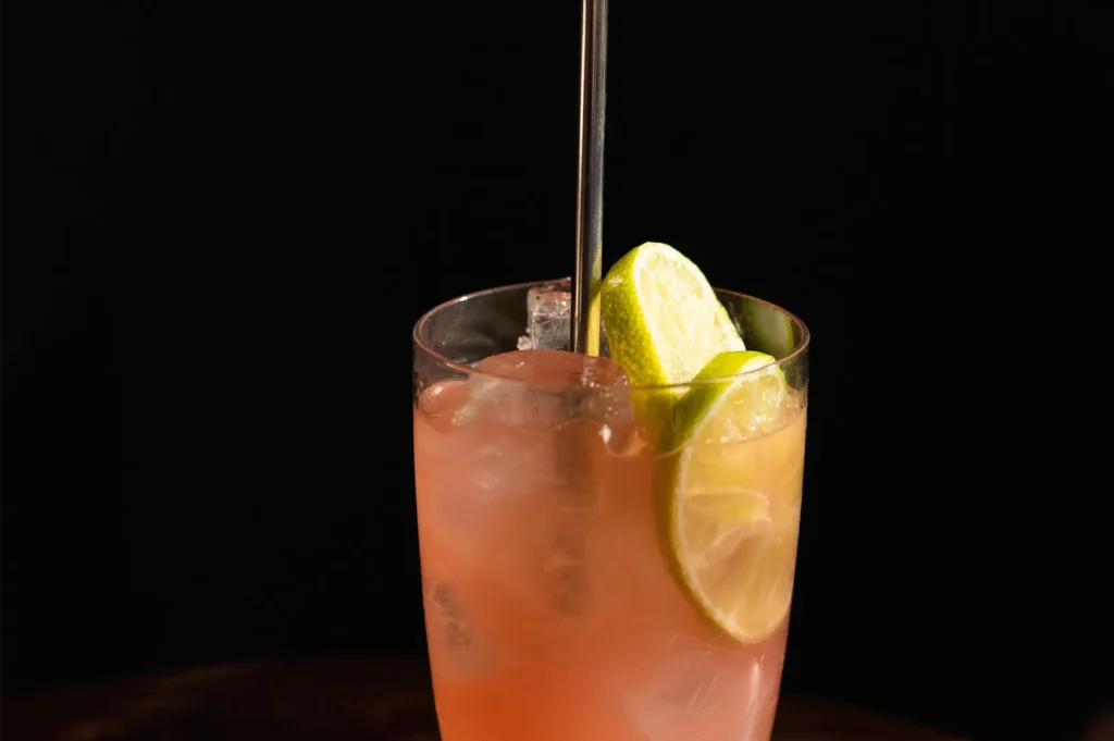 Refreshing pink cocktail in a glass with ice, lime slices, and a metal straw against a dark background.