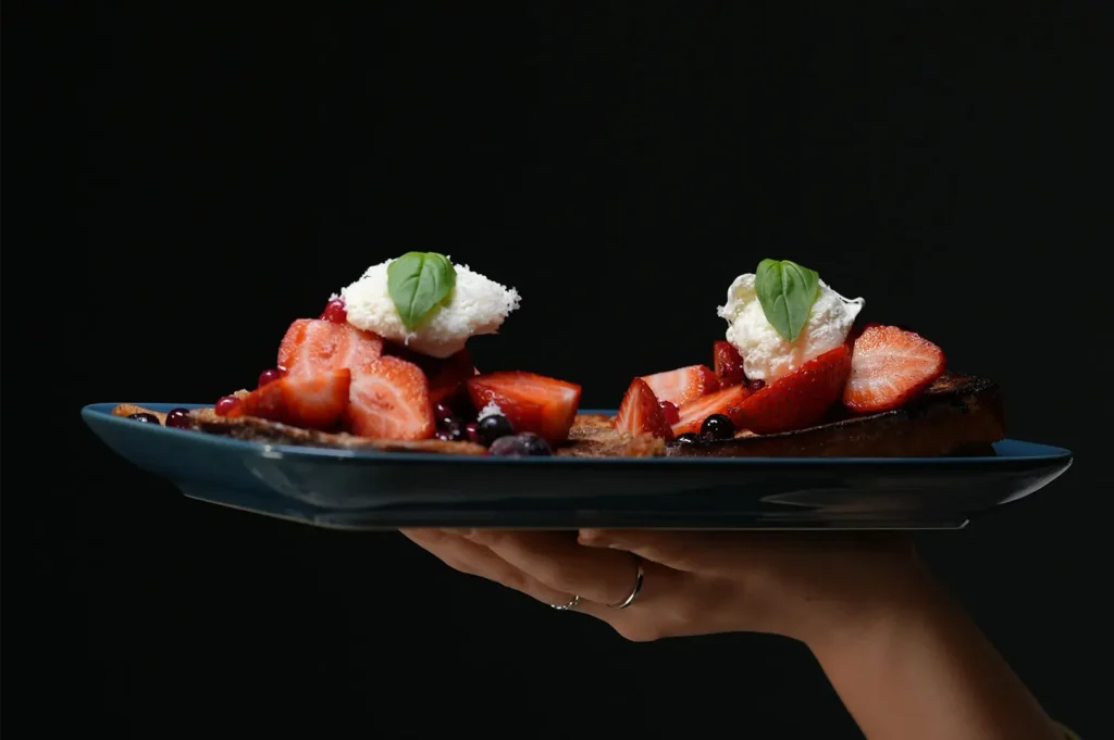 Hand holding a blue plate of french toast topped with fresh strawberries, whipped cream, and basil.