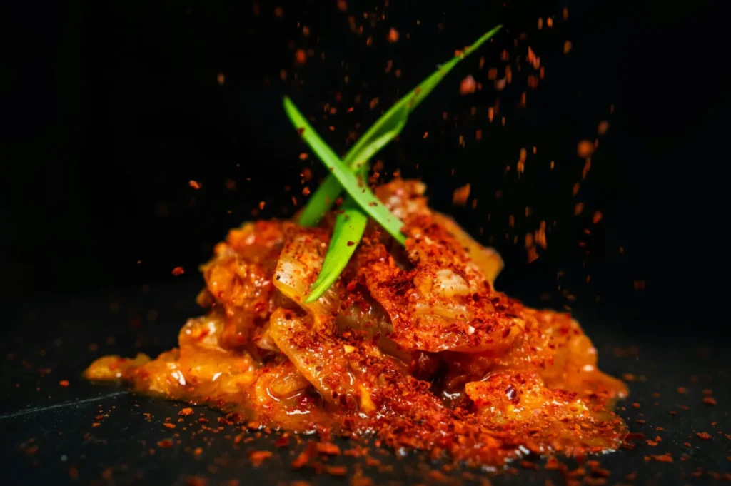 Spicy kimchi dish topped with vibrant red chili flakes and garnished with two green onion stalks, set against a dark background.