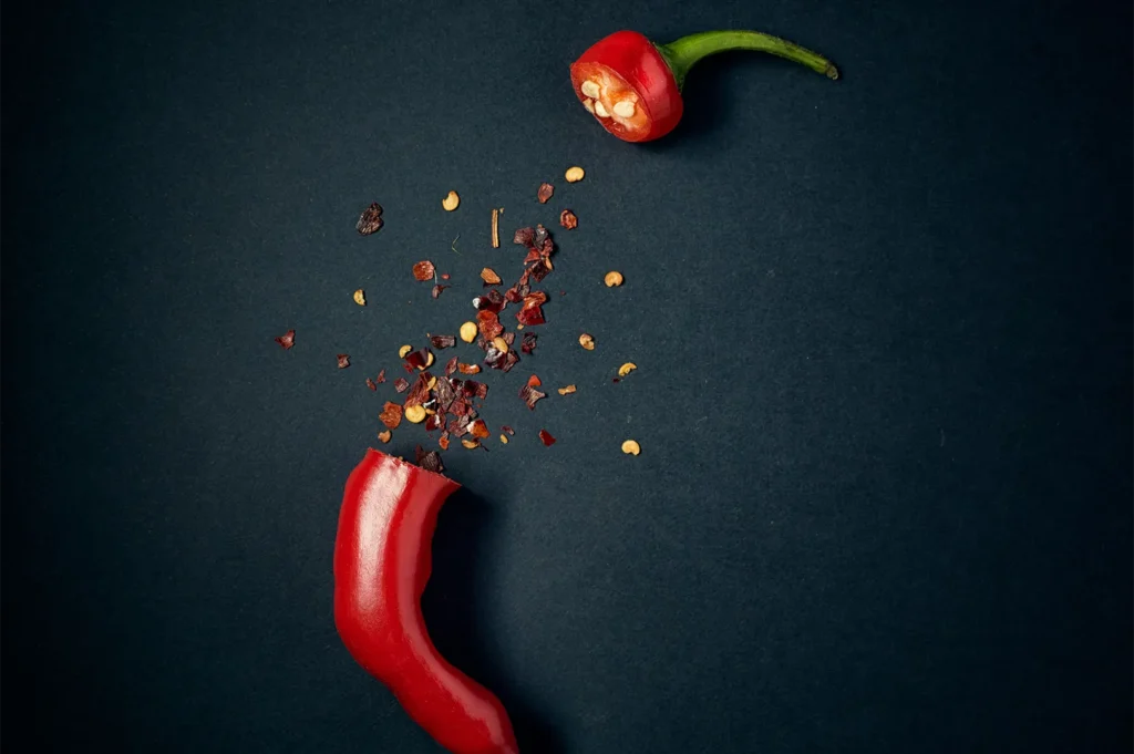 A fresh red chili pepper cut in half with scattered chili flakes and seeds on a dark background.