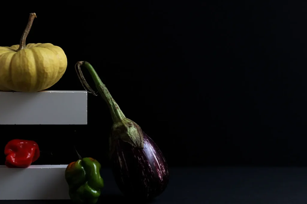 Dark food photography featuring fresh vegetables including a yellow pumpkin, eggplants, and red and green chili peppers arranged against a black background and white blocks.