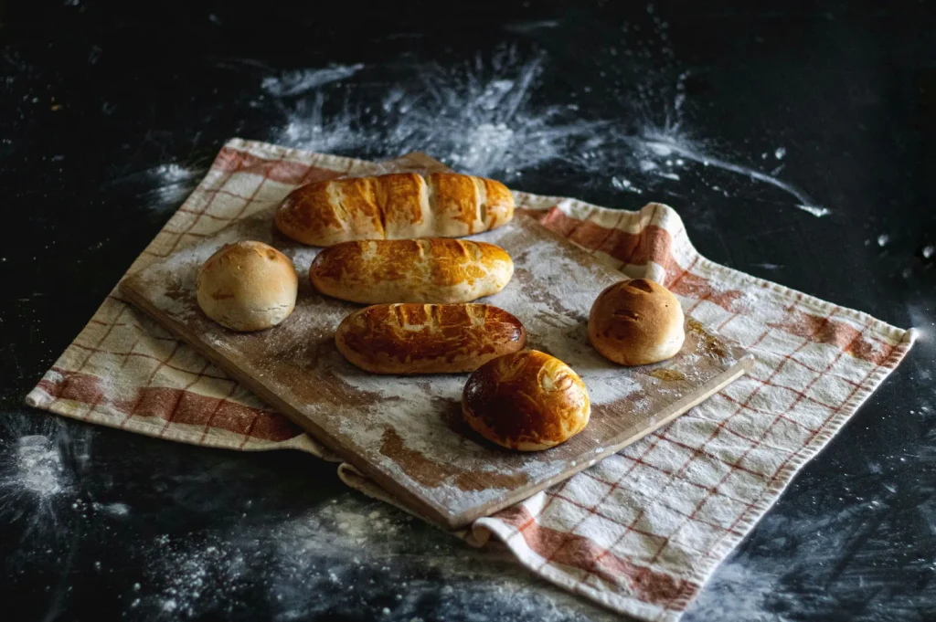 Freshly baked bread rolls and baguettes on a wooden cutting board with a kitchen towel.