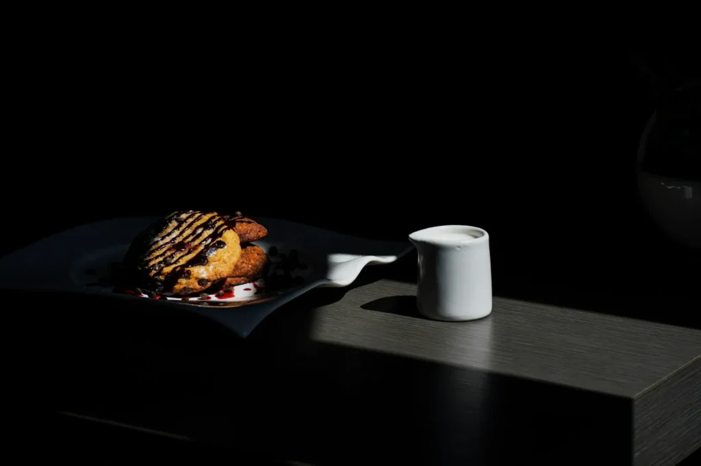 Plate of cookies drizzled with chocolate sauce sits in soft light on a wooden table. Beside it is a small white jug of milk, set against a dark background.