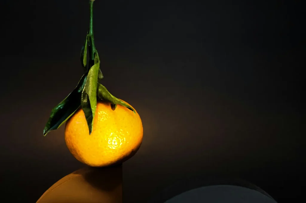 A bright orange tangerine with green leaves balanced on a smooth surface against a dark, minimalist background.