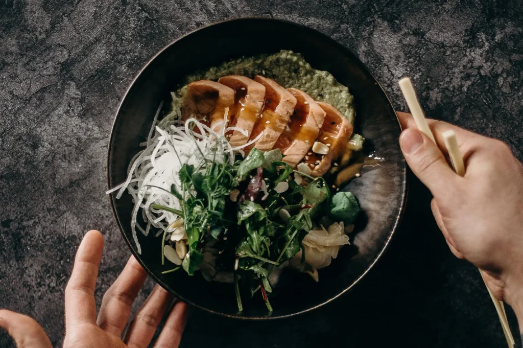 Seared salmon poke bowl with fresh greens and avocado sauce in a dark bowl held with chopsticks.
