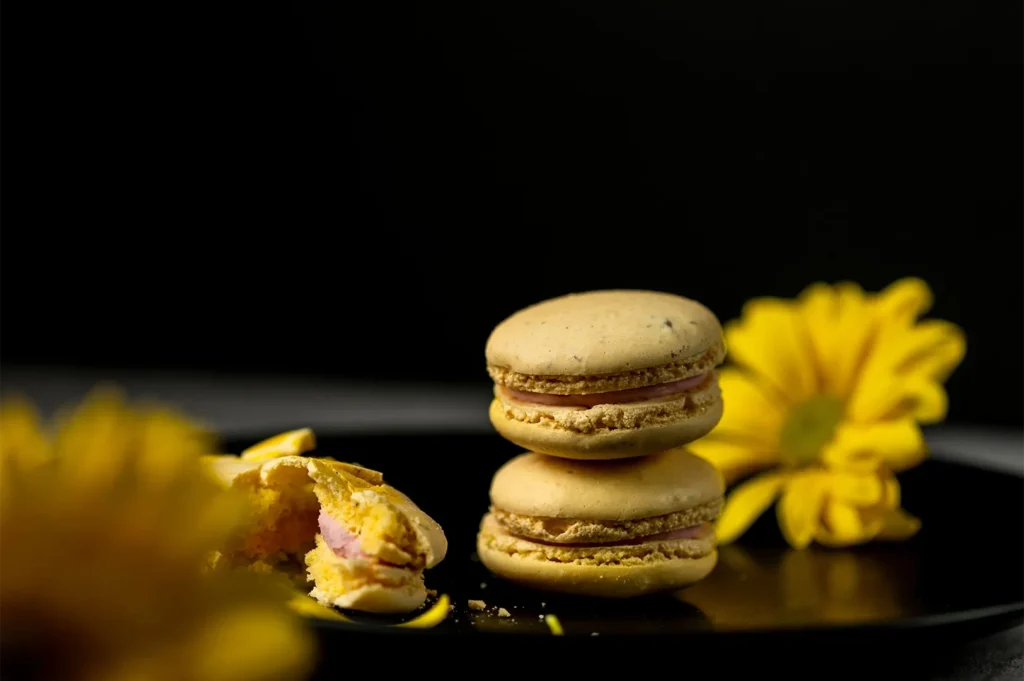 Stack of yellow macarons on a black plate with dramatic low-key rim lighting and yellow flower accents.
