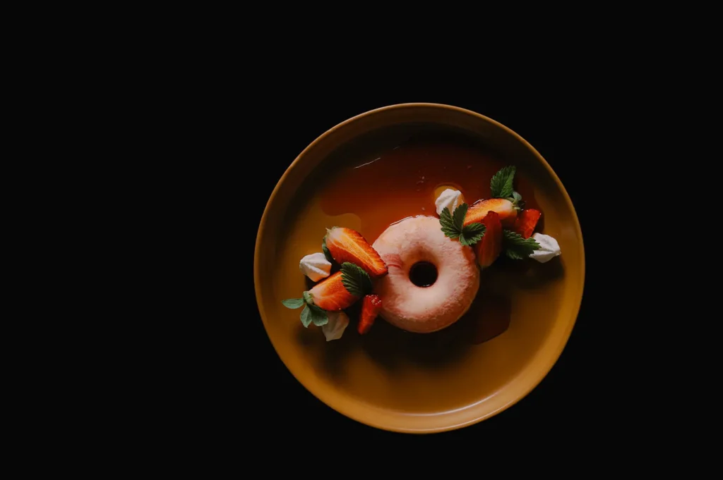 Glazed donut dessert garnished with fresh strawberries, mint, and cream sauce on a yellow plate against a dark background.